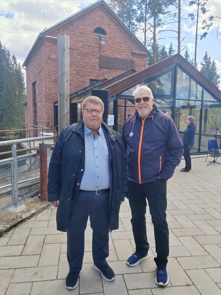 Kuvassa ISU ry.n asiamies Matti Viialainen ja Etelä-Karjalan virkistysaluesäätiön hallituksen puheenjohtaja Ilkka Nokelainen seisovat Kaplan voimalamuseon edustalla vierekkäin ja hymyillen katsovat kameraan. Sää on pilvinen ja kaksikolla on päällään edustavat ulkovaatteet. Matilla on kasvoillaan myös aurinkolasit.